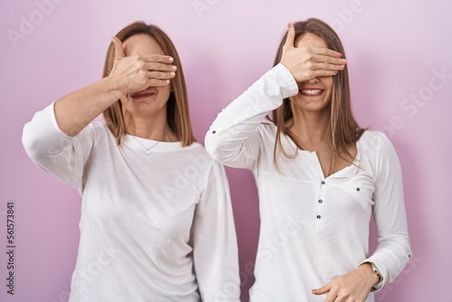 Middle age mother and young daughter standing over pink background smiling and laughing with hand on face covering eyes for surprise. blind concept.