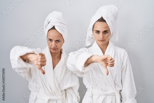 Middle age woman and daughter wearing white bathrobe and towel looking unhappy and angry showing rejection and negative with thumbs down gesture. bad expression.