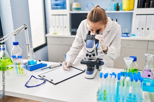 Young blonde woman wearing scientist uniform using microscope working at laboratory