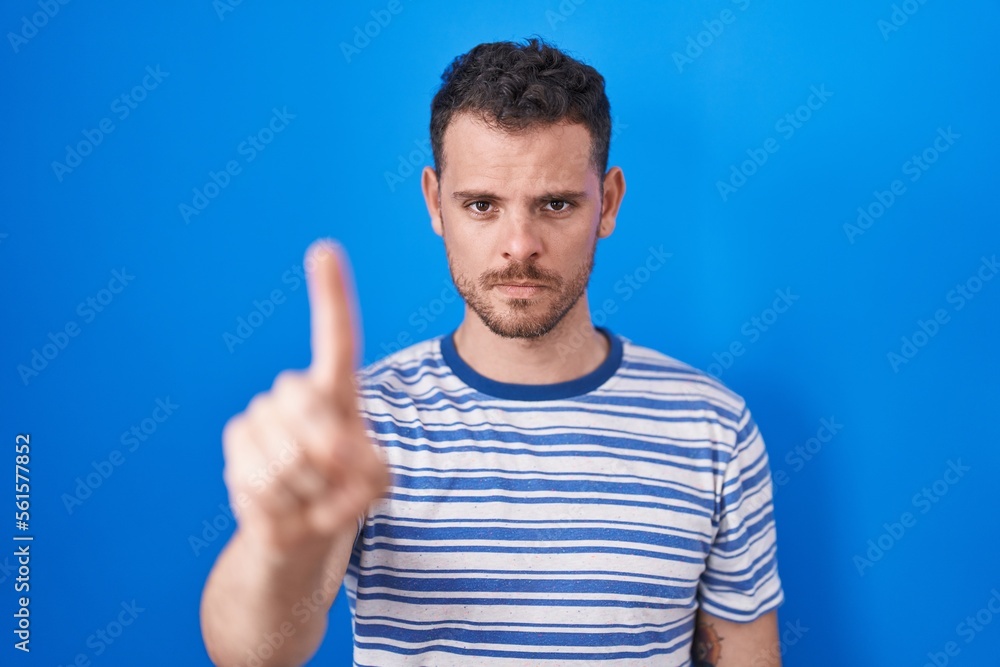 Young hispanic man standing over blue background pointing with finger up and angry expression, showing no gesture Young hispanic man standing over blue background pointing with finger up and angry expression, showing no gesture