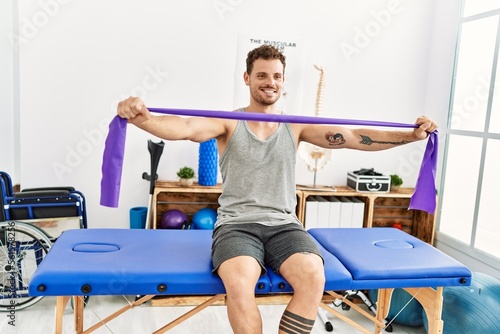 Young hispanic man stretching arm using elastic band sitting on massage board at clinic