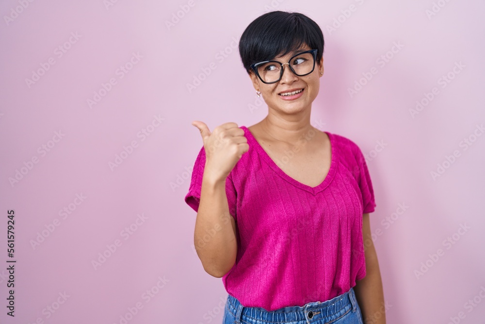 Young asian woman with short hair standing over pink background smiling with happy face looking and pointing to the side with thumb up. Young asian woman with short hair standing over pink background smiling with happy face looking and pointing to the side with thumb up.