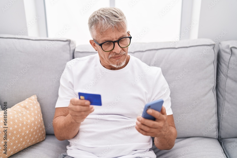 Middle age grey-haired man using smartphone and credit card sitting on sofa at home Middle age grey-haired man using smartphone and credit card sitting on sofa at home