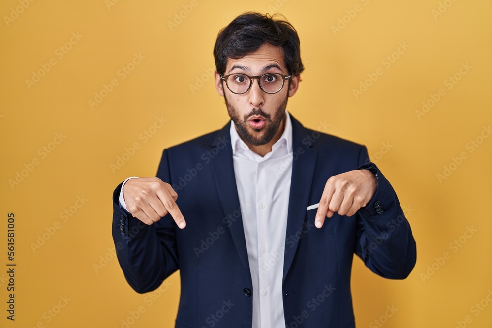 Handsome latin man standing over yellow background pointing down with fingers showing advertisement, surprised face and open mouth Handsome latin man standing over yellow background pointing down with fingers showing advertisement, surprised face and open mouth