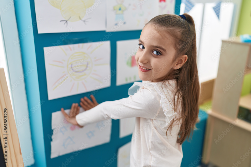 Adorable hispanic girl preschool student hanging draw on wall at kindergarten Adorable hispanic girl preschool student hanging draw on wall at kindergarten