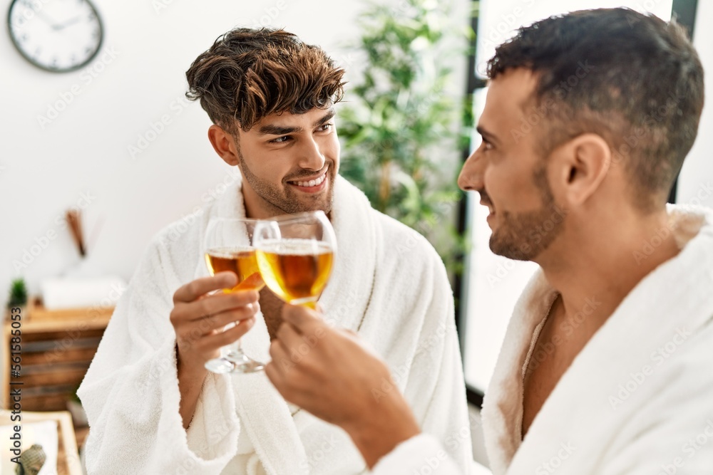 Two hispanic men couple toasting with champagne sitting on massage table at beauty center