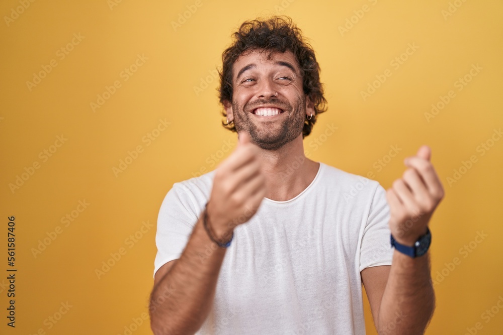 Young hispanic man smiling confident doing spend money gesture over isolated yellow background Young hispanic man smiling confident doing spend money gesture over isolated yellow background
