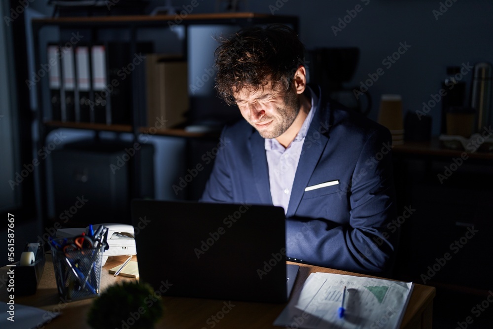 Hispanic young man working at the office at night with hand on stomach because nausea, painful disease feeling unwell. ache concept. Hispanic young man working at the office at night with hand on stomach because nausea, painful disease feeling unwell. ache concept.