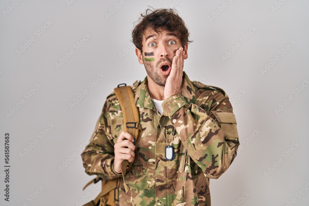 Hispanic young man wearing camouflage army uniform afraid and shocked, surprise and amazed expression with hands on face