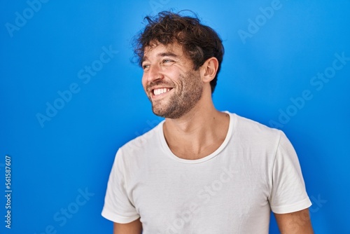Hispanic young man standing over blue background looking away to side with smile on face, natural expression. laughing confident.