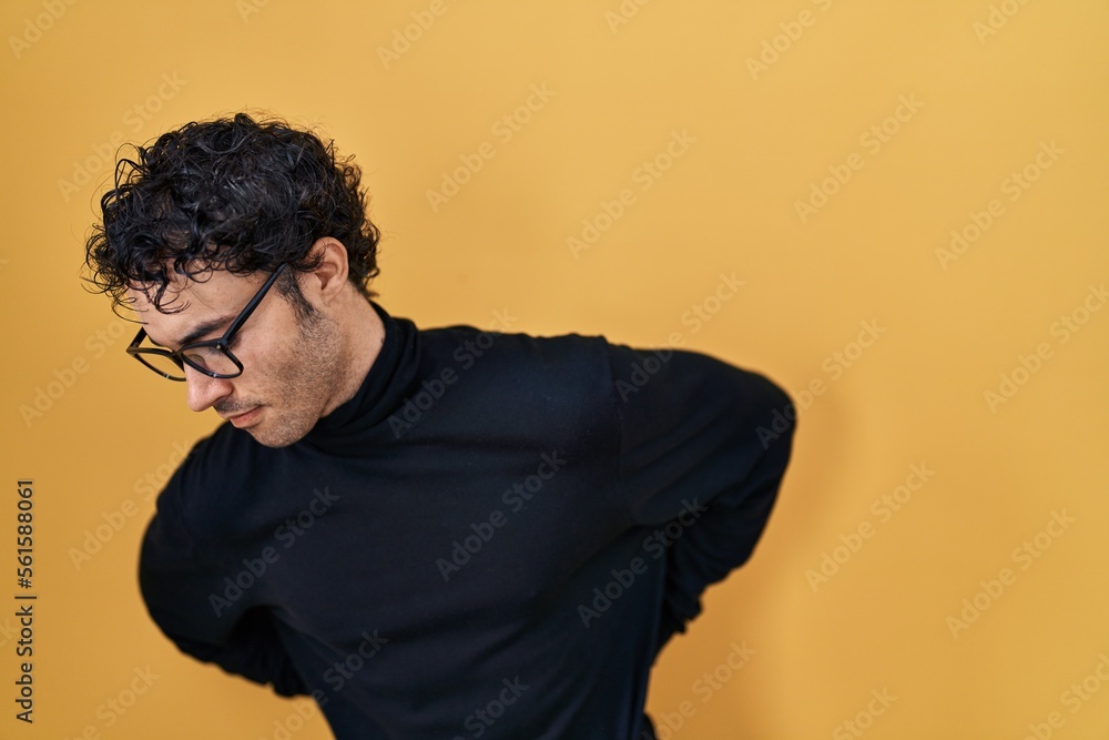Hispanic man standing over yellow background suffering of backache, touching back with hand, muscular pain Hispanic man standing over yellow background suffering of backache, touching back with hand, muscular pain