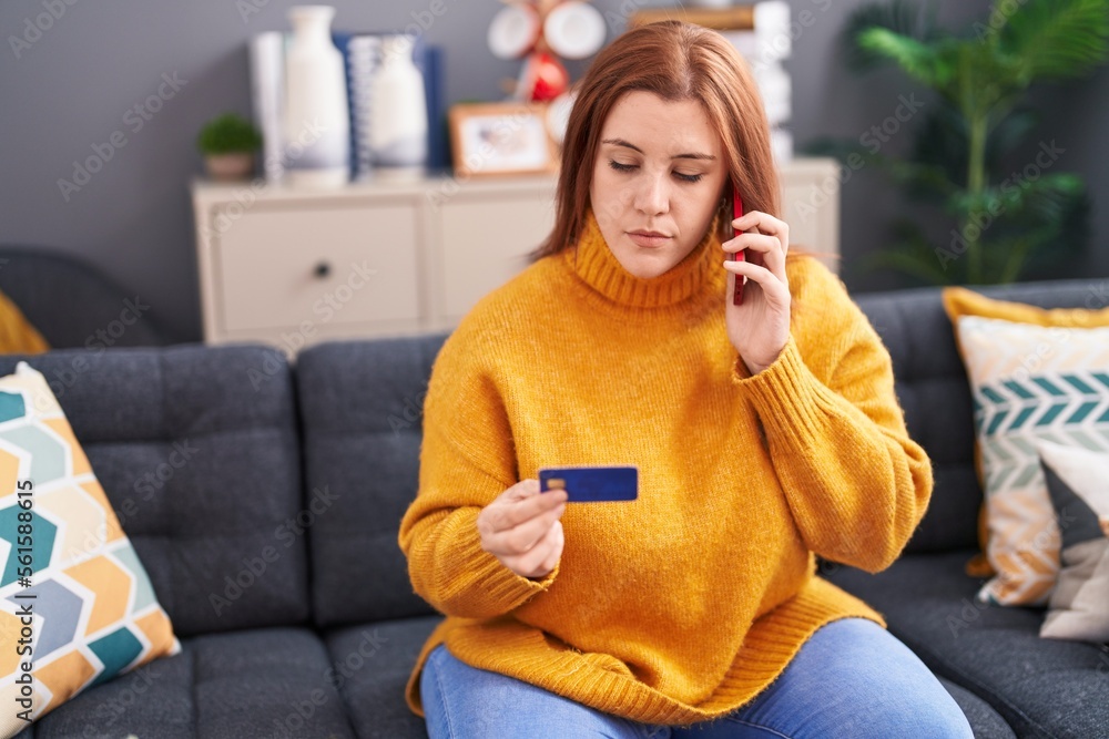 Young beautiful plus size woman talking on smartphone holding credit card with serious expression at home Young beautiful plus size woman talking on smartphone holding credit card with serious expression at home