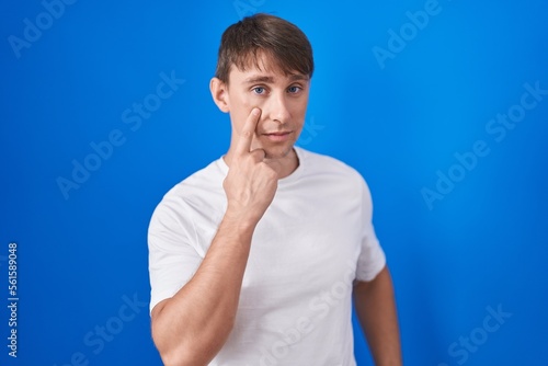 Caucasian blond man standing over blue background pointing to the eye watching you gesture, suspicious expression