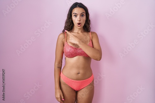 Young hispanic woman wearing lingerie over pink background surprised pointing with finger to the side, open mouth amazed expression.
