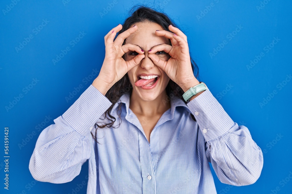 Young brunette woman standing over blue background doing ok gesture like binoculars sticking tongue out, eyes looking through fingers. crazy expression. Young brunette woman standing over blue background doing ok gesture like binoculars sticking tongue out, eyes looking through fingers. crazy expression.