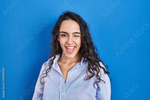 Young brunette woman standing over blue background winking looking at the camera with sexy expression, cheerful and happy face.