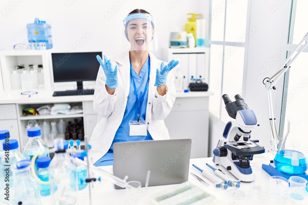 Young hispanic woman wearing scientist uniform crazy and mad shouting and yelling with aggressive expression and arms raised. frustration concept. Young hispanic woman wearing scientist uniform crazy and mad shouting and yelling with aggressive expression and arms raised. frustration concept.