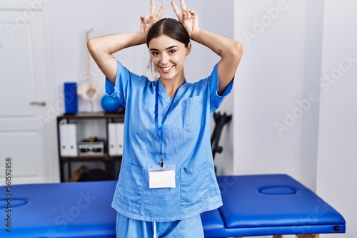 Young hispanic woman wearing physiotherapist uniform standing at clinic posing funny and crazy with fingers on head as bunny ears  smiling cheerful