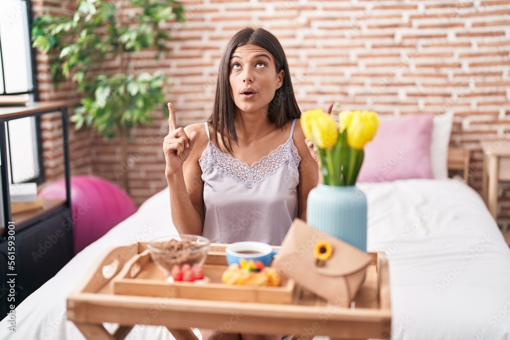 Brunette young woman eating breakfast sitting on the bed amazed and surprised looking up and pointing with fingers and raised arms.