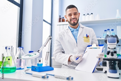 Hispanic man working at scientist laboratory holding blank clipboard clueless and confused expression. doubt concept.
