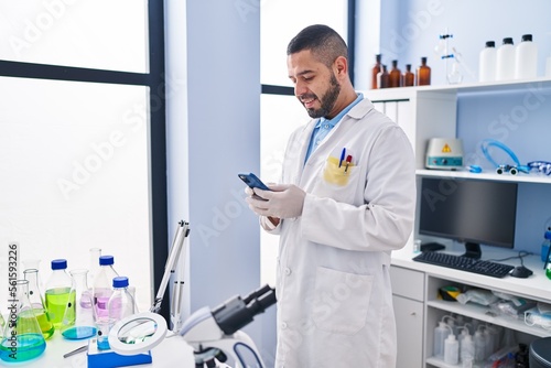 Young latin man scientist smiling confident using smartphone at laboratory