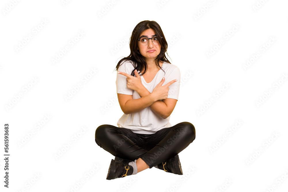 Young indian woman sitting on the floor cut out isolated points sideways, is trying to choose between two options. Young indian woman sitting on the floor cut out isolated points sideways, is trying to choose between two options.