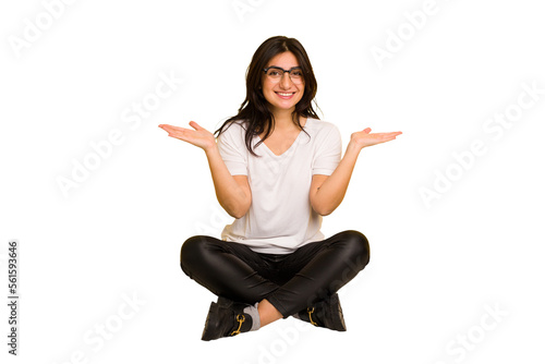 Young indian woman sitting on the floor cut out isolated makes scale with arms, feels happy and confident. © Asier