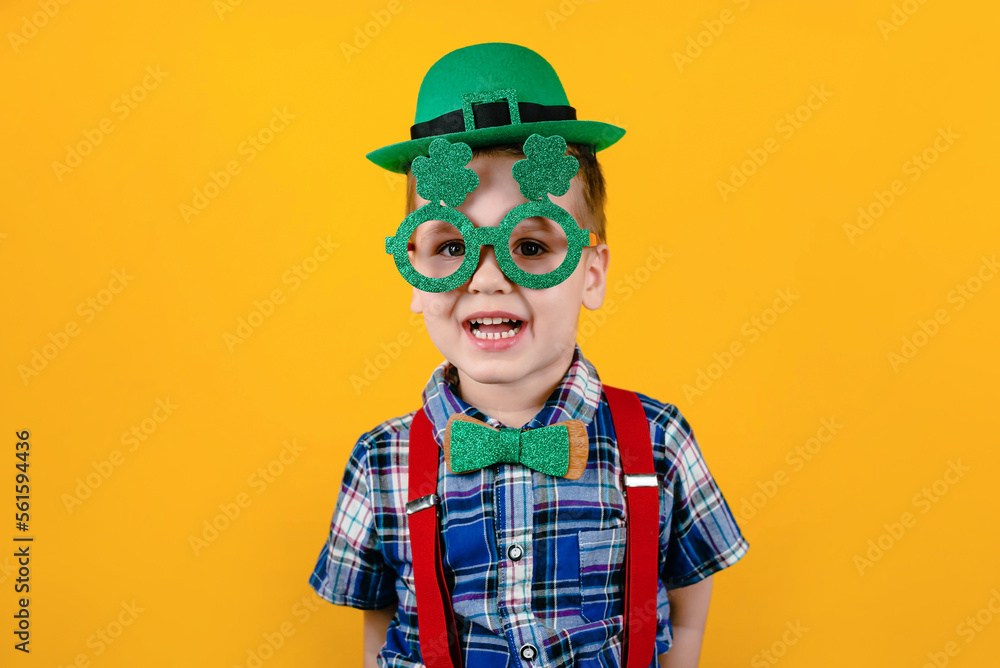 Fototapeta premium Funny toddler boy with green glasses and a hat on a yellow background. Happy Patrick's Day.