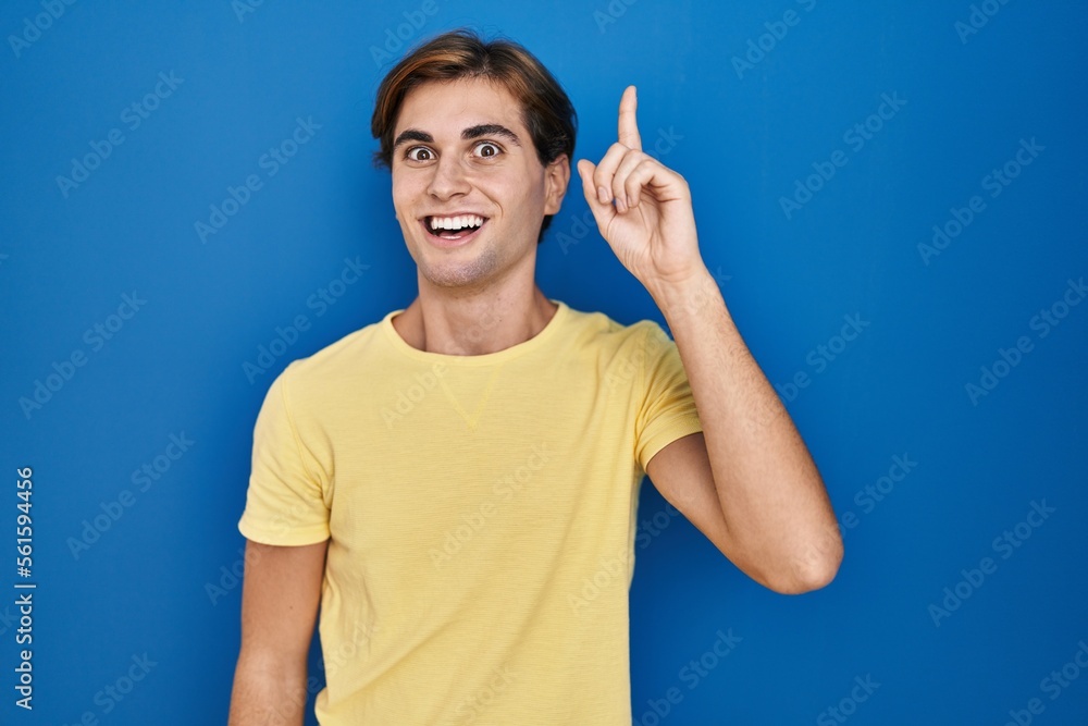 Young man standing over blue background pointing finger up with successful idea. exited and happy. number one. Young man standing over blue background pointing finger up with successful idea. exited and happy. number one.