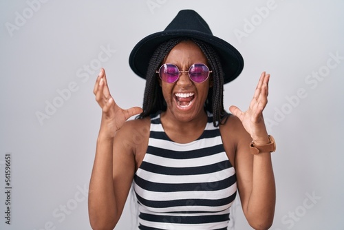 Young african american with braids wearing hat and sunglasses celebrating mad and crazy for success with arms raised and closed eyes screaming excited. winner concept