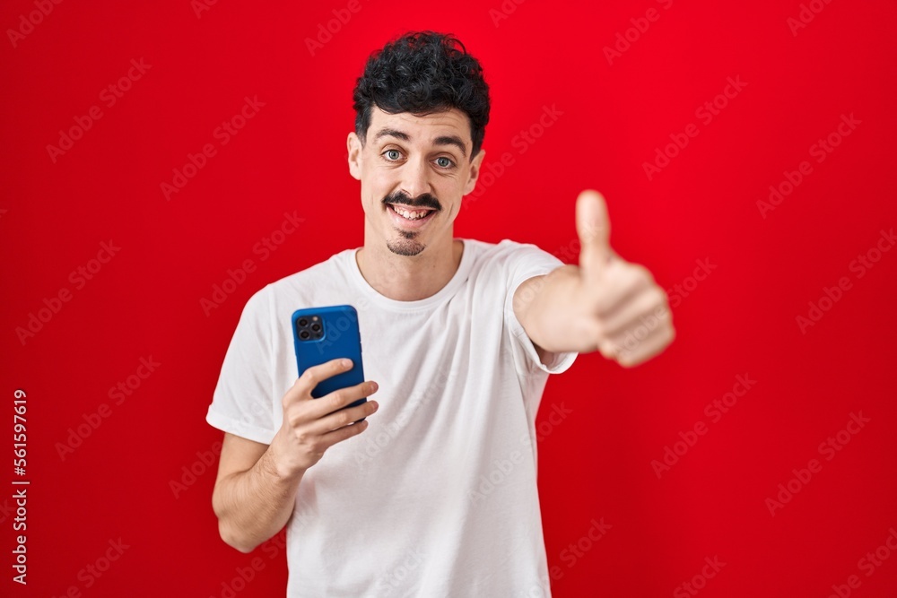 Hispanic man using smartphone over red background approving doing positive gesture with hand, thumbs up smiling and happy for success. winner gesture. Hispanic man using smartphone over red background approving doing positive gesture with hand, thumbs up smiling and happy for success. winner gesture.