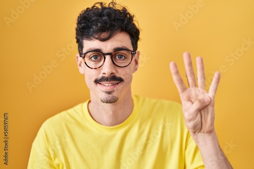 Hispanic man wearing glasses standing over yellow background showing and pointing up with fingers number four while smiling confident and happy.