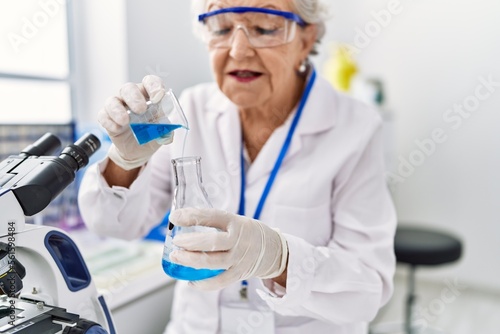 Senior grey-haired woman wearing scientist uniform pouring liquid at laboratory