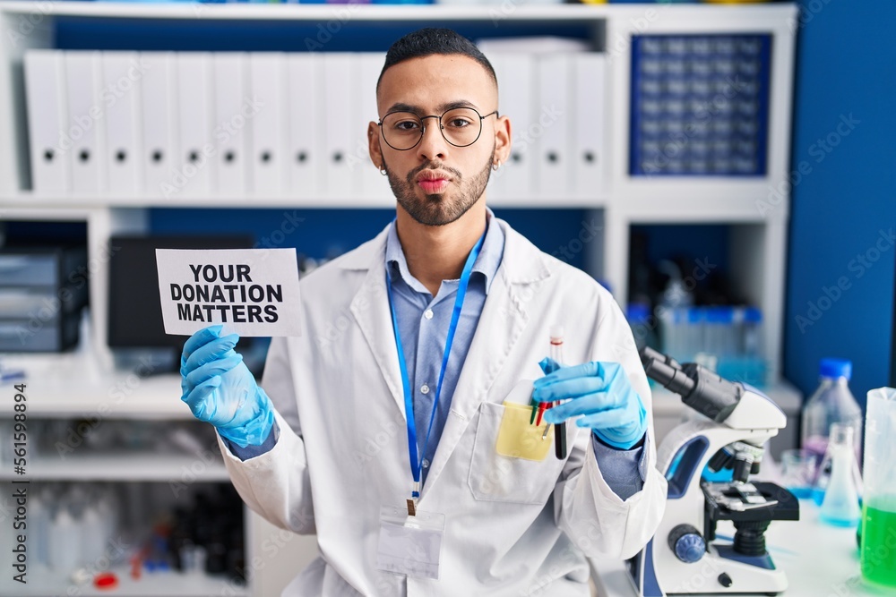 Young hispanic man working at scientist laboratory holding your donation matters holding blood sample looking at the camera blowing a kiss being lovely and sexy. love expression.