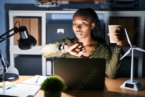 African woman working using computer laptop at night checking the time on wrist watch, relaxed and confident