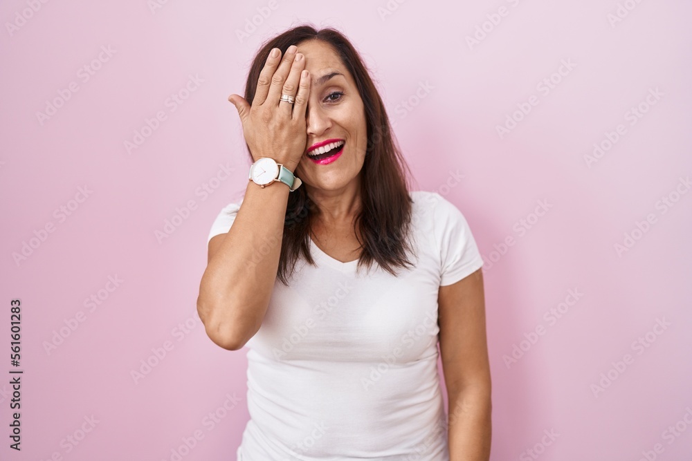 Middle age brunette woman standing over pink background covering one eye with hand, confident smile on face and surprise emotion. Middle age brunette woman standing over pink background covering one eye with hand, confident smile on face and surprise emotion.