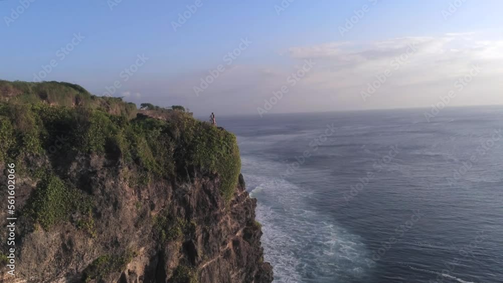 Loving couple of people is staying on the edge of the cliff above ocean on beautiful sunset. Romantic atmosphere, aerial view, drone shot. Uluwatu area in Bali, Indonesia Loving couple of people is staying on the edge of the cliff above ocean on beautiful sunset. Romantic atmosphere, aerial view, drone shot. Uluwatu area in Bali, Indonesia