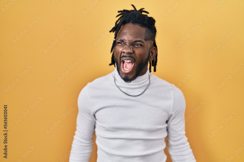 African man with dreadlocks wearing turtleneck sweater over yellow background angry and mad screaming frustrated and furious, shouting with anger. rage and aggressive concept. African man with dreadlocks wearing turtleneck sweater over yellow background angry and mad screaming frustrated and furious, shouting with anger. rage and aggressive concept.