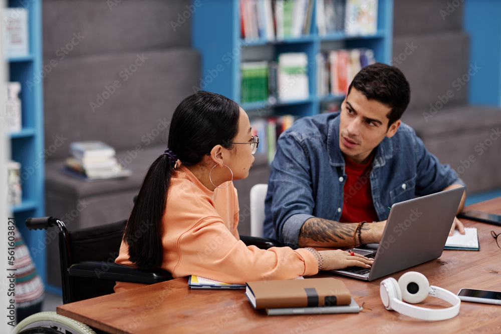 Fototapeta premium Teenage girl in wheelchair studying with friend in college library