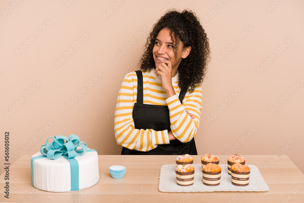 Young african american woman preparing a sweet cake and muffins on a table relaxed thinking about something looking at a copy space. Young african american woman preparing a sweet cake and muffins on a table relaxed thinking about something looking at a copy space.