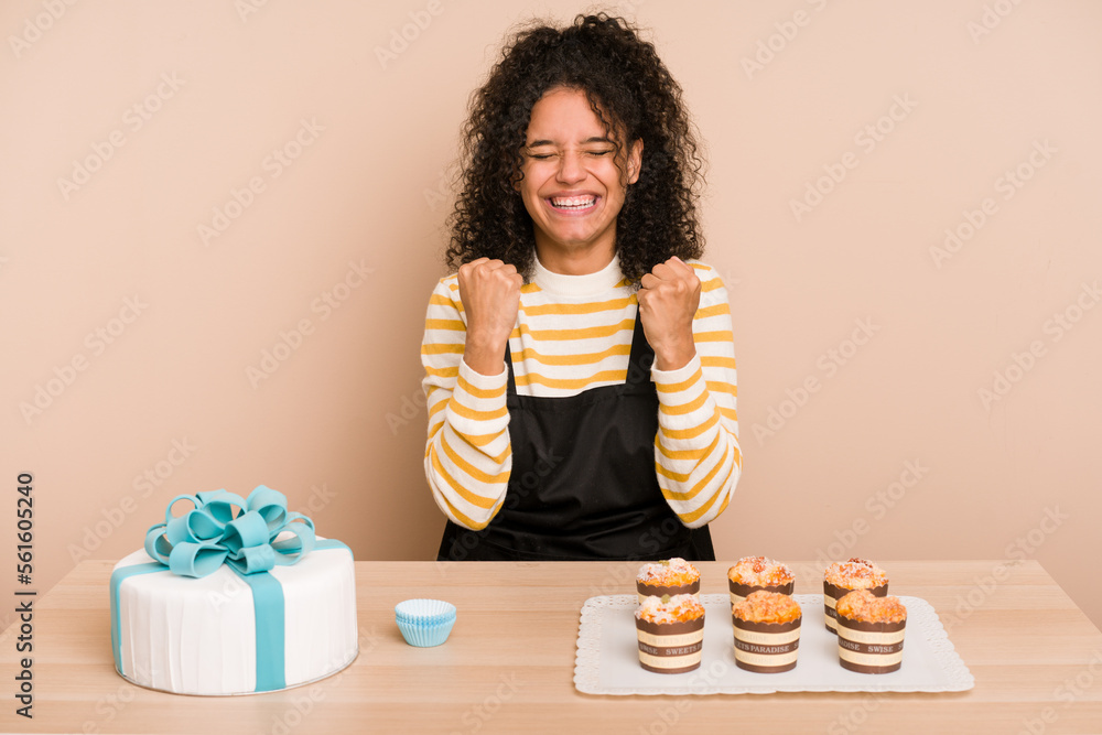 Young african american woman preparing a sweet cake and muffins on a table cheering carefree and excited. Victory concept. Young african american woman preparing a sweet cake and muffins on a table cheering carefree and excited. Victory concept.