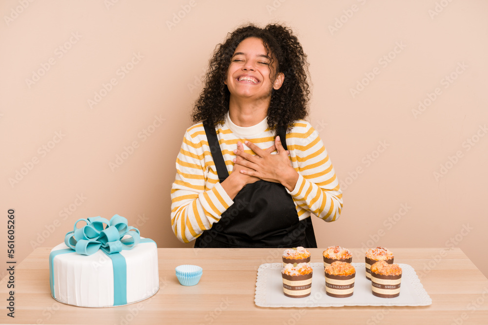 Young african american woman preparing a sweet cake and muffins on a table laughing keeping hands on heart, concept of happiness.
