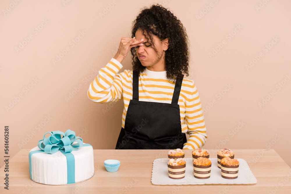 Young african american woman preparing a sweet cake and muffins on a table having a head ache, touching front of the face. Young african american woman preparing a sweet cake and muffins on a table having a head ache, touching front of the face.