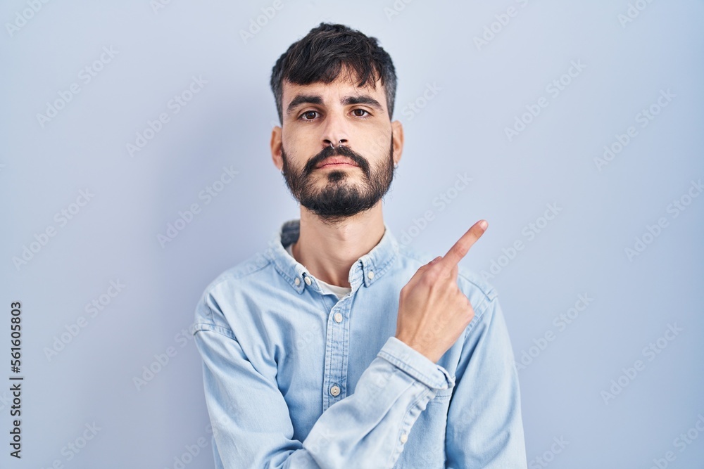 Young hispanic man with beard standing over blue background pointing with hand finger to the side showing advertisement, serious and calm face Young hispanic man with beard standing over blue background pointing with hand finger to the side showing advertisement, serious and calm face