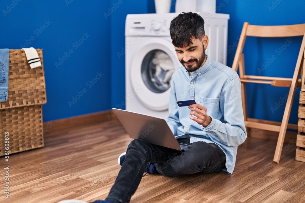 Young hispanic man using laptop and credit card waiting for washing machine at laundry room Young hispanic man using laptop and credit card waiting for washing machine at laundry room