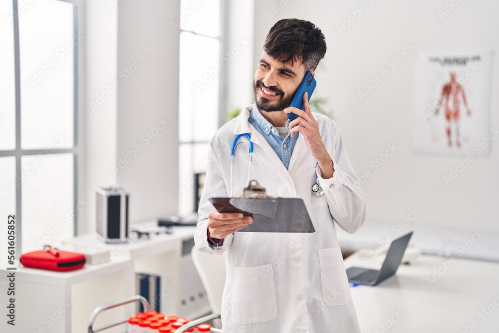Young hispanic man wearing doctor uniform talking on smartphone reading medical report at clinic Young hispanic man wearing doctor uniform talking on smartphone reading medical report at clinic