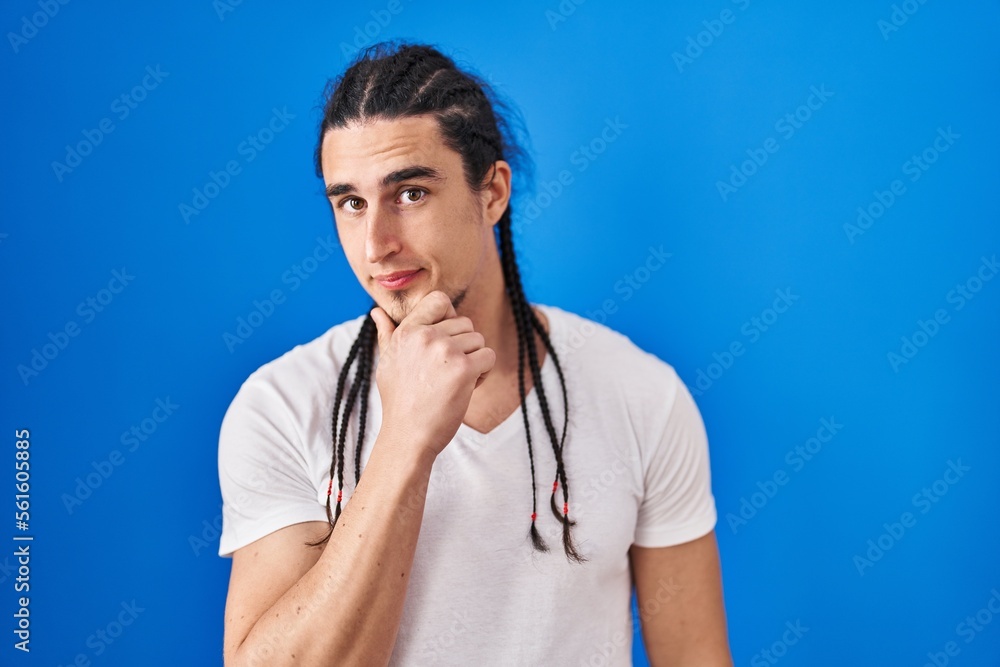 Hispanic man with long hair standing over blue background looking confident at the camera with smile with crossed arms and hand raised on chin. thinking positive. Hispanic man with long hair standing over blue background looking confident at the camera with smile with crossed arms and hand raised on chin. thinking positive.