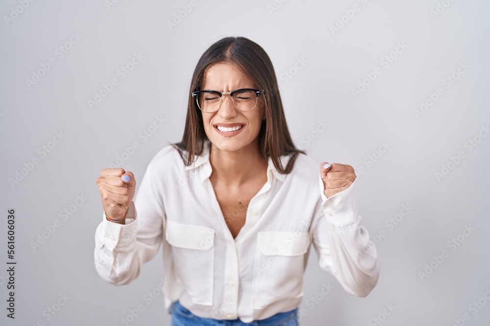 Young brunette woman wearing glasses angry and mad raising fists frustrated and furious while shouting with anger. rage and aggressive concept. Young brunette woman wearing glasses angry and mad raising fists frustrated and furious while shouting with anger. rage and aggressive concept.