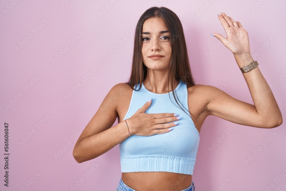Young brunette woman standing over pink background swearing with hand on chest and open palm, making a loyalty promise oath