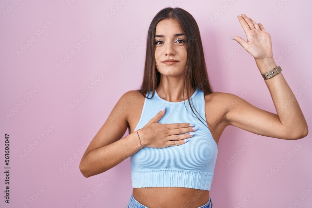 Young brunette woman standing over pink background swearing with hand on chest and open palm, making a loyalty promise oath Young brunette woman standing over pink background swearing with hand on chest and open palm, making a loyalty promise oath